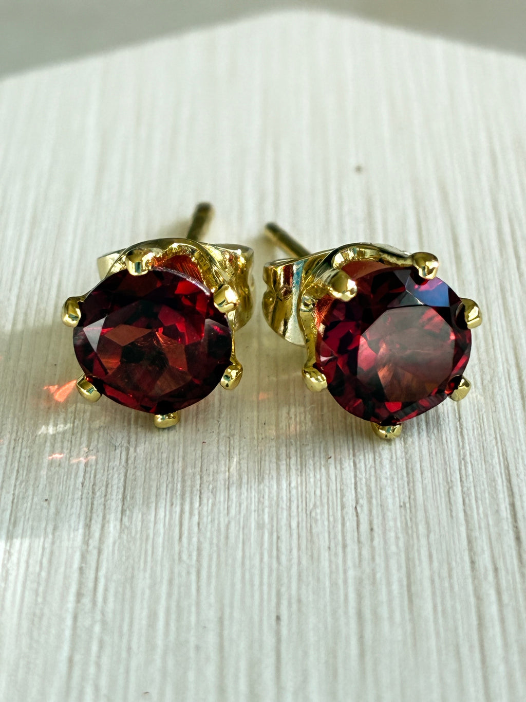 Pair of garnet gemstone stud earrings with gold settings on a light wooden surface.
