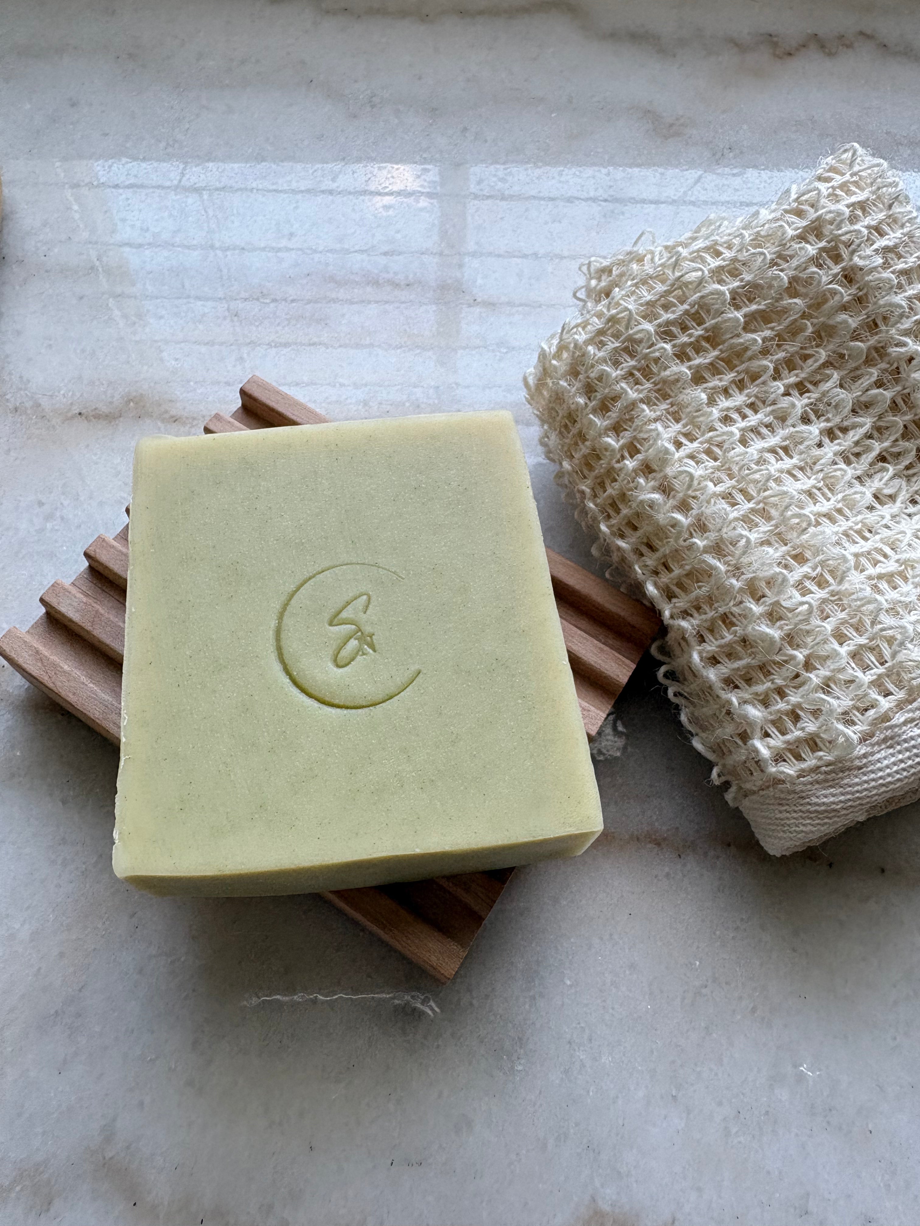 A cucumber soap resting on a wooden soap dish next to a beige woven scrubber on a light marble surface.