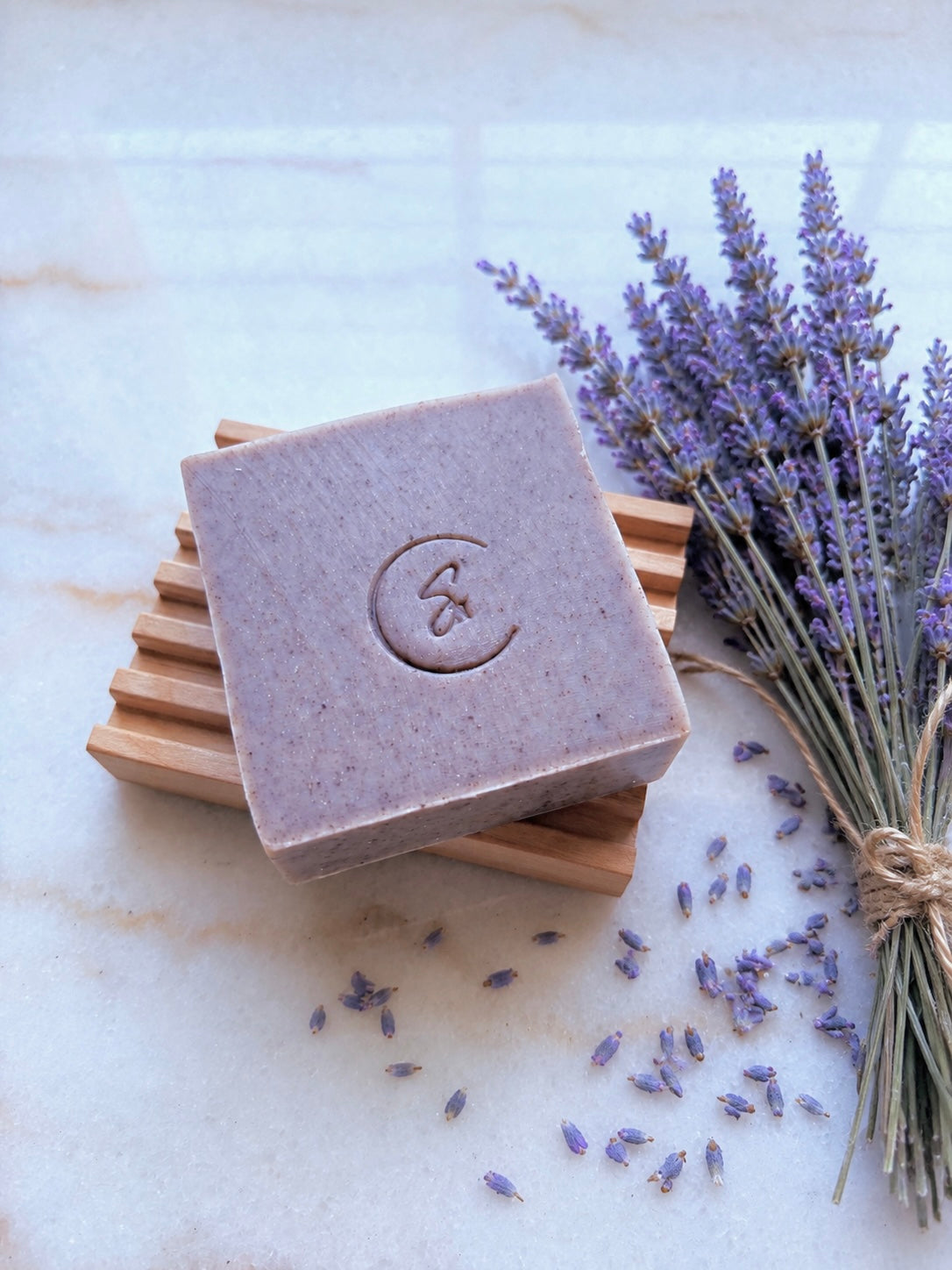 A rectangular lavender soap bar with resting on a wooden soap dish beside a bundle of dried lavender sprigs on a light marble surface.