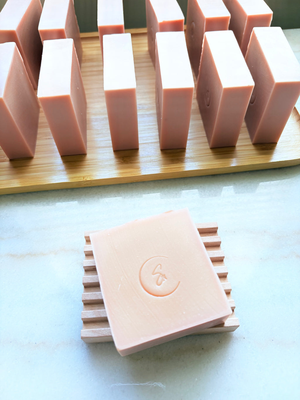 A pale pink rectangular bar of soap with an embossed crescent moon and stylized symbol, resting on a wooden soap dish in the foreground, with multiple matching soap bars standing on a wooden tray in the background.