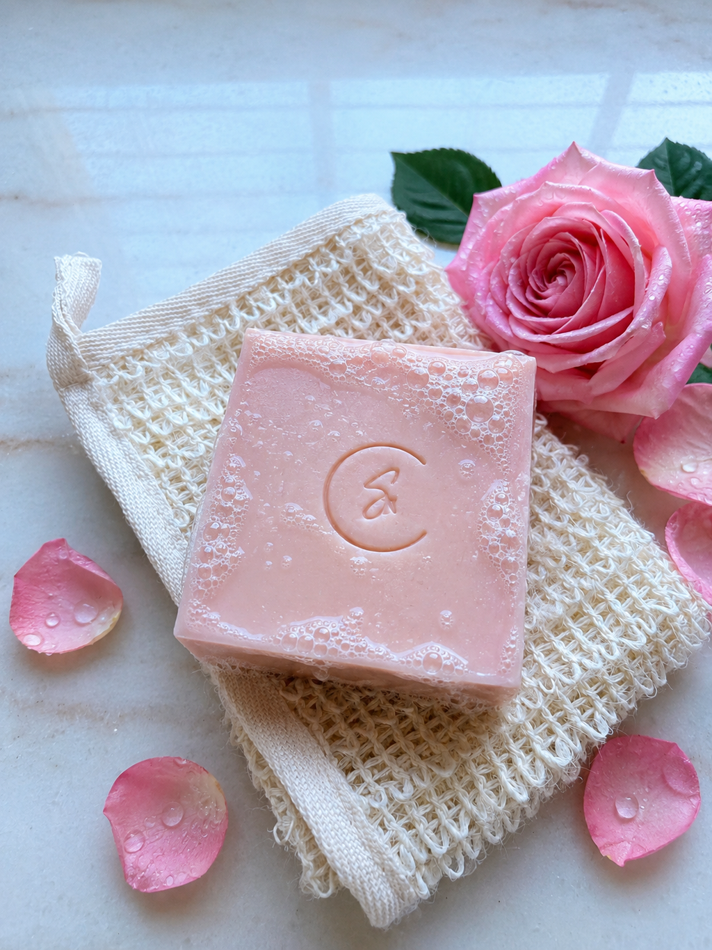 A Rose soap resting on a cream-colored mesh soap bag. The setup is styled with a fresh pink rose, scattered pink petals, and water droplets on a light marble surface.