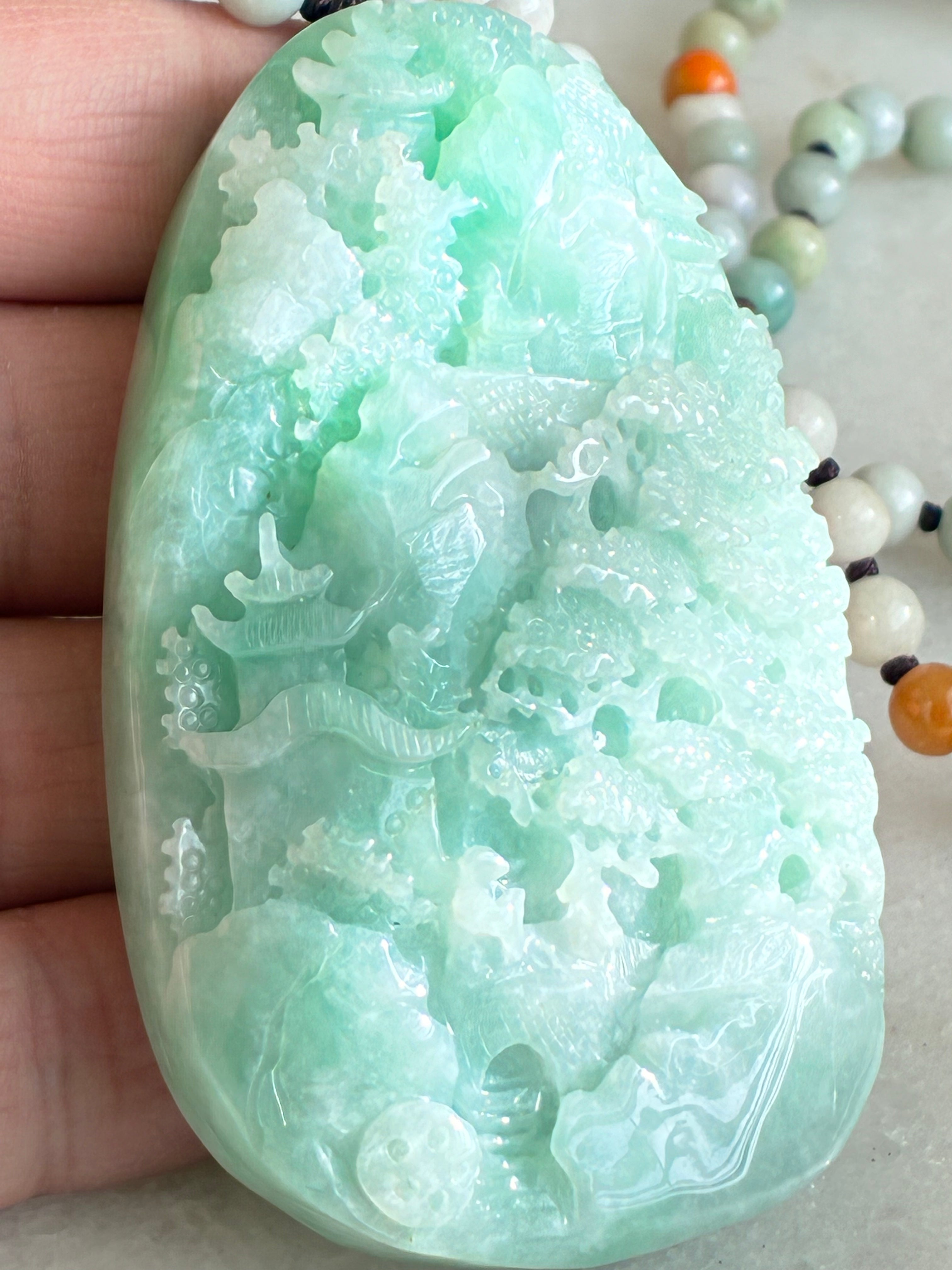 Green jade pendant with intricate carvings held by a hand, with beads in the background.